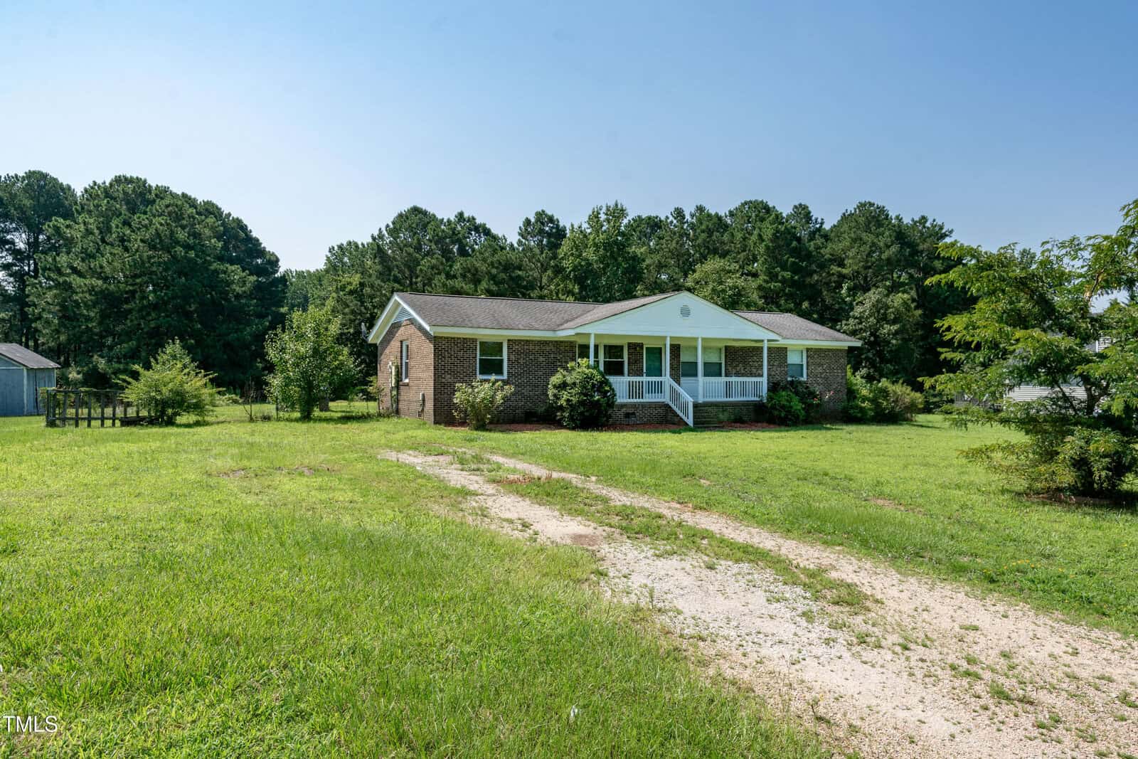 A single-story brick house with a white front porch sits on a large grassy lot with a gravel driveway, surrounded by trees under a clear blue sky.