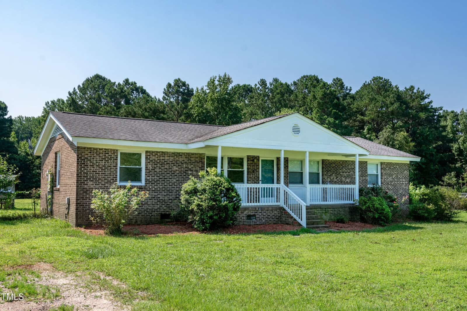A one-story brick house with a covered front porch, white columns, and surrounding shrubs, set on a grassy lawn with trees in the background under a clear blue sky.