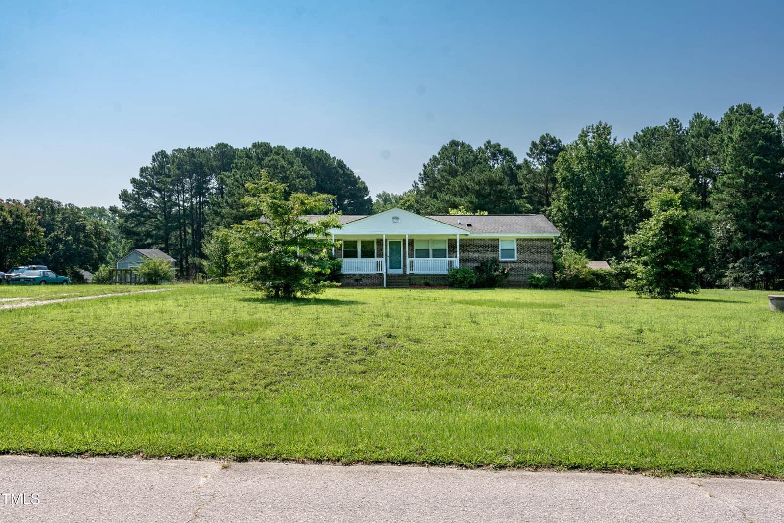 A single-story brick house with a white porch sits on a large, grassy lawn bordered by trees under a clear blue sky. The house is set back from the street with a wide expanse of grass in front.