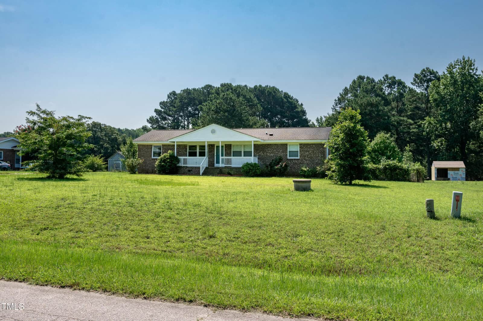 A single-story brick house with white columns on the porch sits on a large, grassy lawn with a few small trees. There are woods in the background and a shed to the right. The sky is clear and blue.