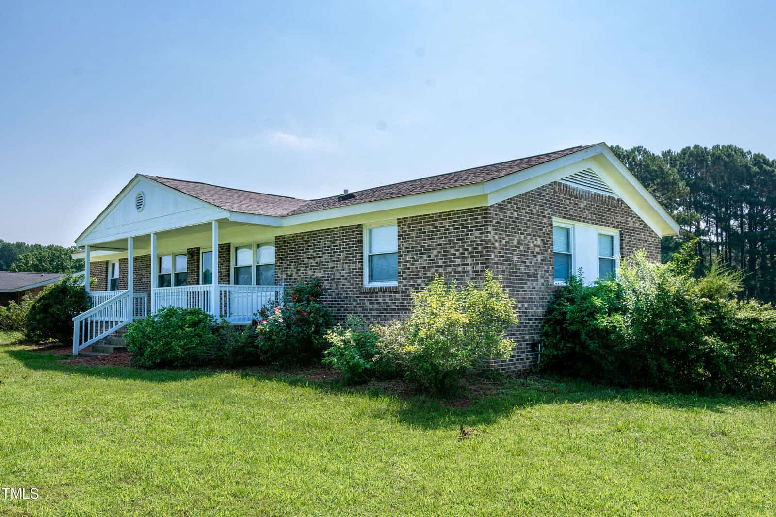 One-story brick house with a covered front porch and white railing, surrounded by green grass and shrubs, with trees in the background under a clear blue sky.