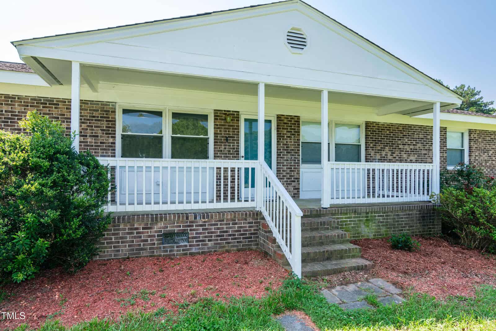 Front view of a single-story brick house with a white porch, railing, and three steps leading to the entrance. Two windows and a white door are visible. Mulch and shrubs are in the foreground. Sky is clear.