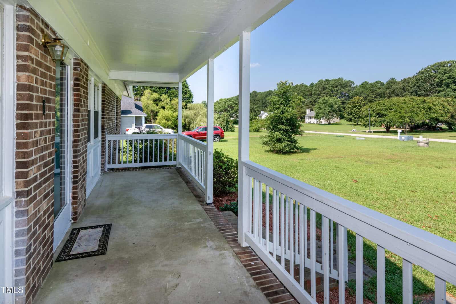 Covered front porch with white railing, brick accents, and concrete floor. A welcome mat is by the door. The porch overlooks a grassy yard with trees and neighboring houses visible in the background.