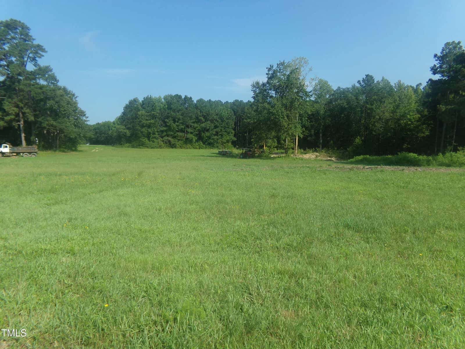 A wide grassy field with scattered trees along the edge, under a clear blue sky. A small trailer is visible on the left side near the trees.