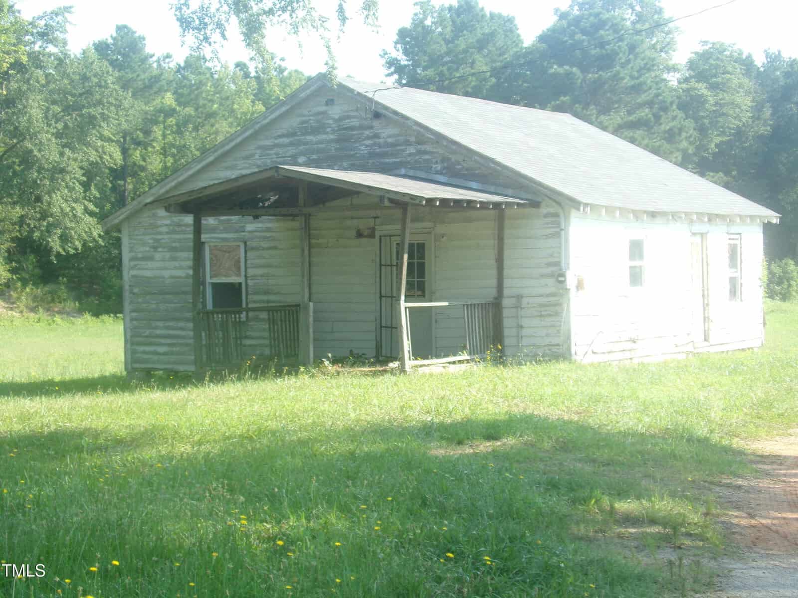 A small, weathered, white wooden house with a damaged porch, missing railings, and peeling paint sits on a grassy lot with trees in the background under a sunny sky.