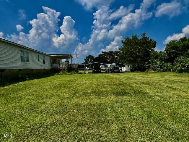 A large, neatly mowed grassy yard leads to a white manufactured home on the left, with several cars parked under metal carports in the background, and a partly cloudy blue sky overhead.