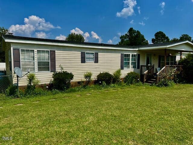 Located at 174 Daffodil Drive in Henderson, this single-story manufactured home features beige siding, dark shutters, a covered front porch, several windows, a satellite dish, and bushes along the foundation in a grassy yard.