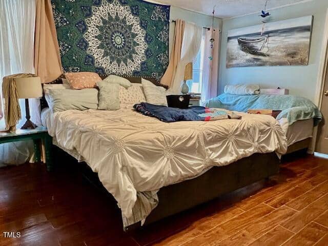 Bedroom with two beds, one large and one small. The large bed has white bedding and decorative pillows, with a blue tapestry on the wall behind it. Wood floors, curtains, and a painting of a boat are visible.