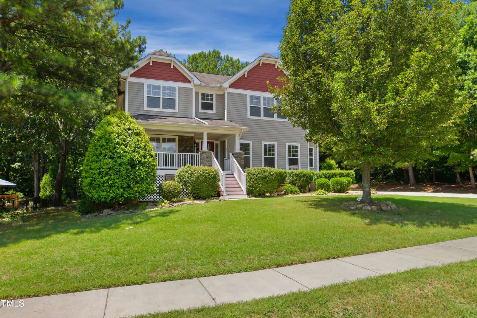 Two-story suburban house with gray siding, red upper accents, white trim, a covered front porch, and steps. Green lawn, manicured bushes, mature trees, and a sidewalk in front. Clear blue sky overhead.