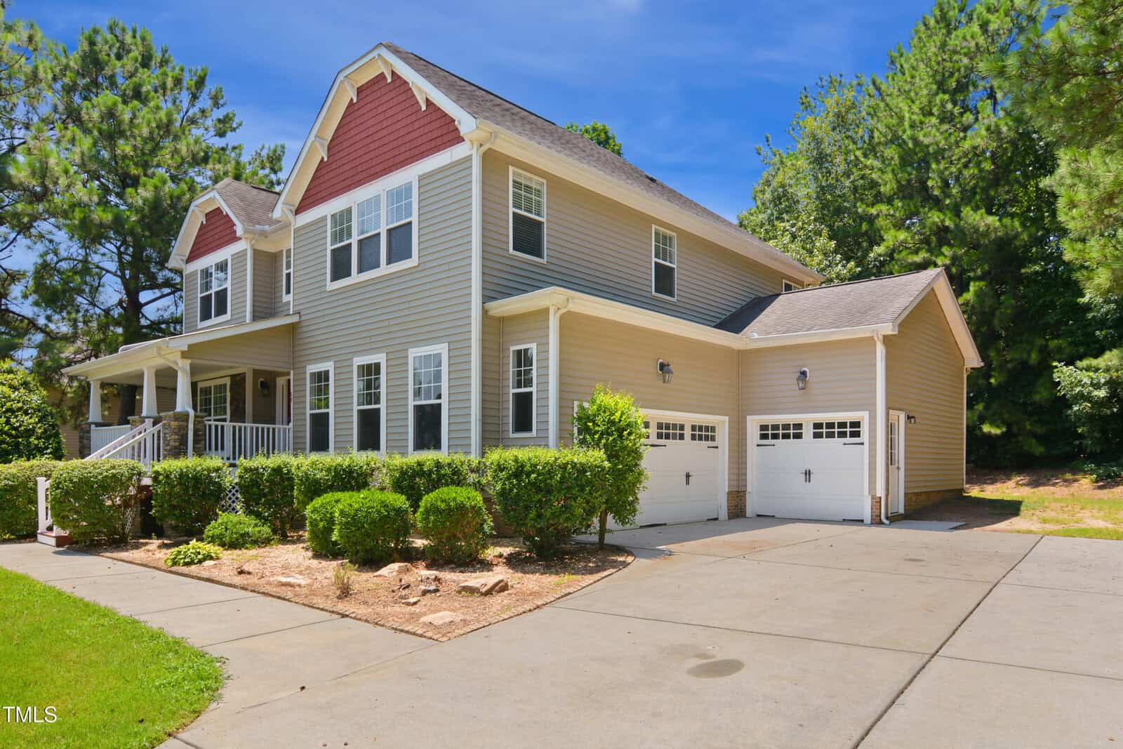 Two-story house with gray siding, white trim, and a red accent above the porch. The home features a covered front porch, two-car garage, neatly trimmed bushes, and a wide concrete driveway. Trees and grass surround the property.