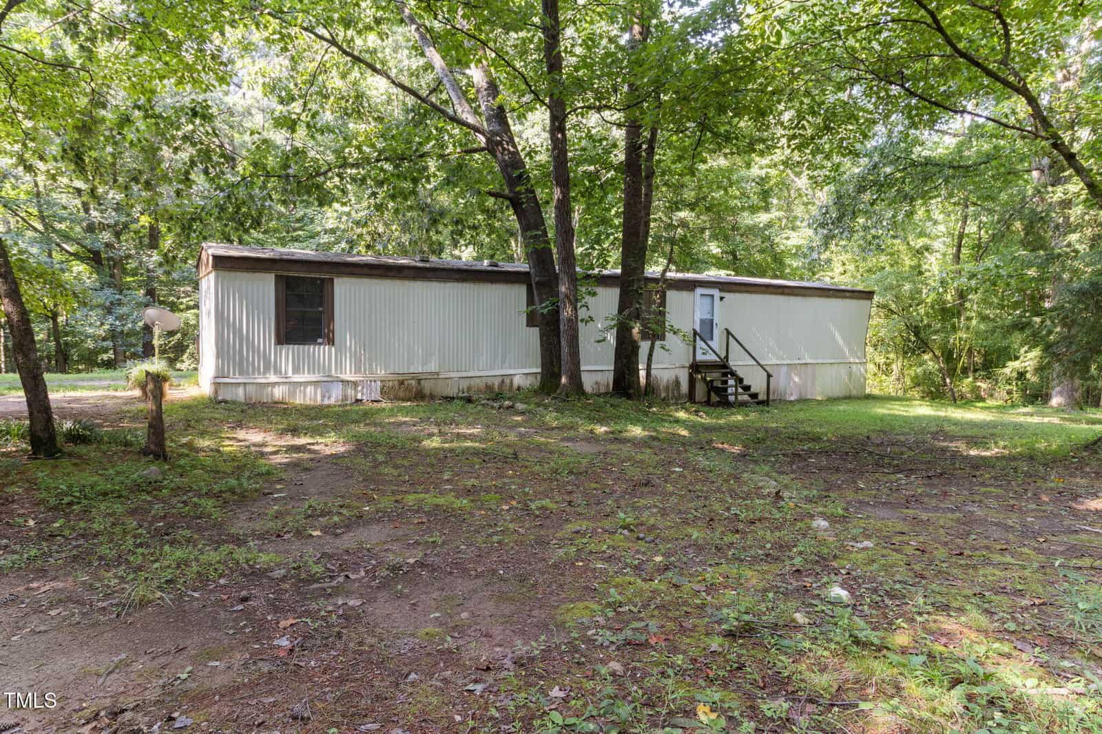 A white mobile home at 3170 Loblolly Lane sits in a wooded area of Wake Forest, with grass and trees surrounding it; the ground is patchy with dirt and leaves and a small set of stairs leads to the front door.