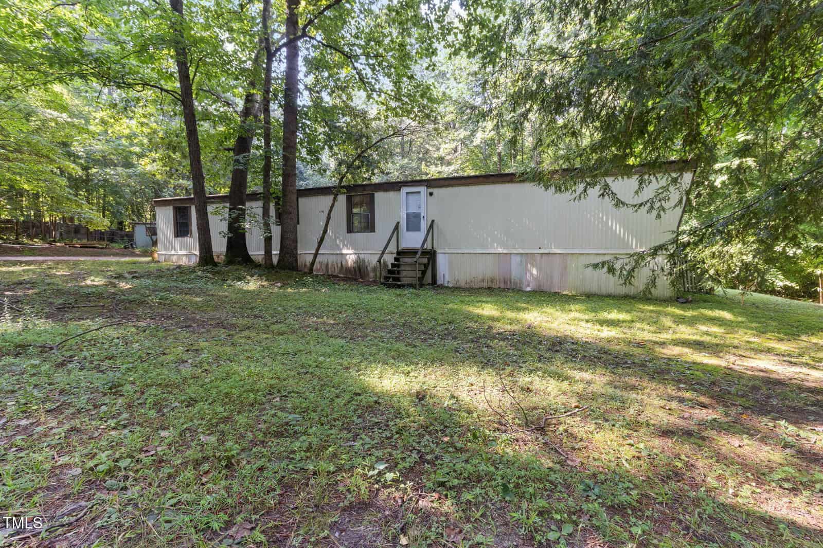 A beige mobile home with a white door and small front steps sits on a grassy, shaded lot surrounded by tall trees. Sunlight filters through the leaves, creating a dappled effect on the lawn and home.