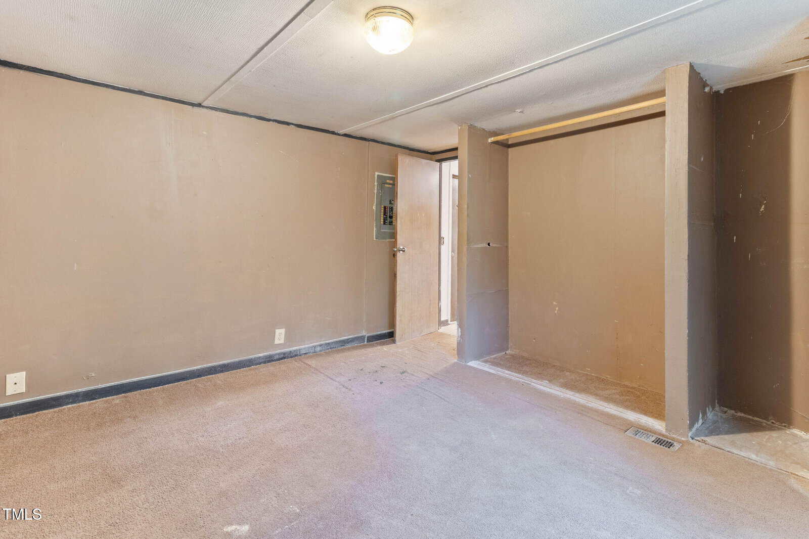 Empty room with beige carpet, brown walls, and exposed electrical panel. There is an open closet area without doors and a single ceiling light fixture. A small window is partially visible near the doorway.