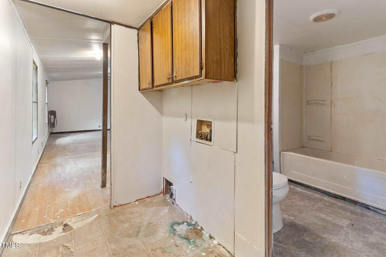 A hallway with worn floors leads to a room in the distance. An open area shows laundry hookups below wooden cabinets and a view into a bathroom with a toilet and bathtub. Surfaces appear unfinished or in need of repair.