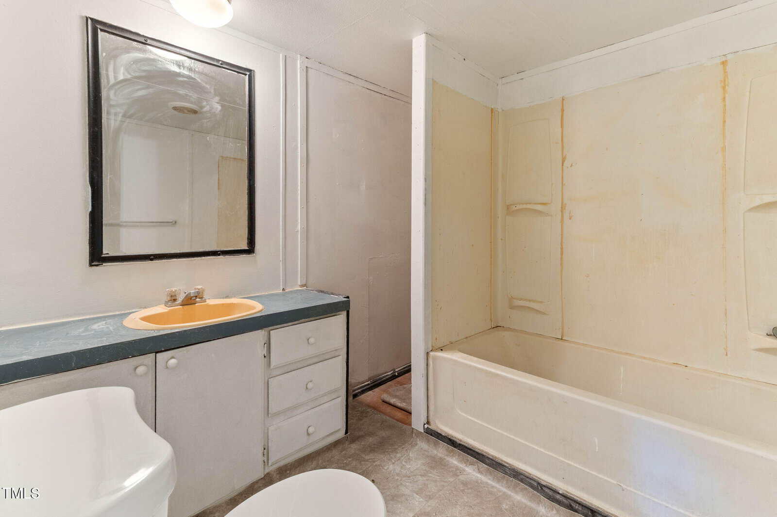 A bathroom with a yellow sink set in a blue countertop, a wall-mounted mirror, a bathtub with aged or stained walls, a toilet, and a light fixture on the ceiling. The walls and floor appear worn.