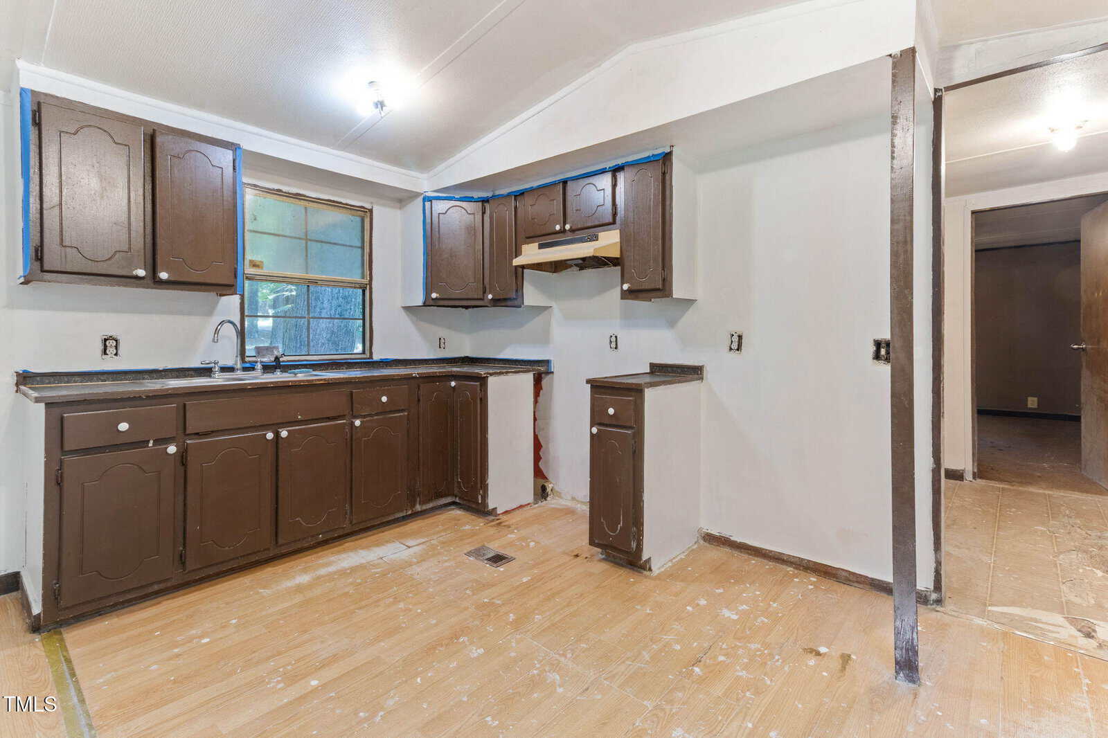 A partially renovated kitchen with brown cabinets, a black countertop, an empty space for an appliance, exposed outlets, and unfinished flooring. A doorway leads to another room with visible construction materials.