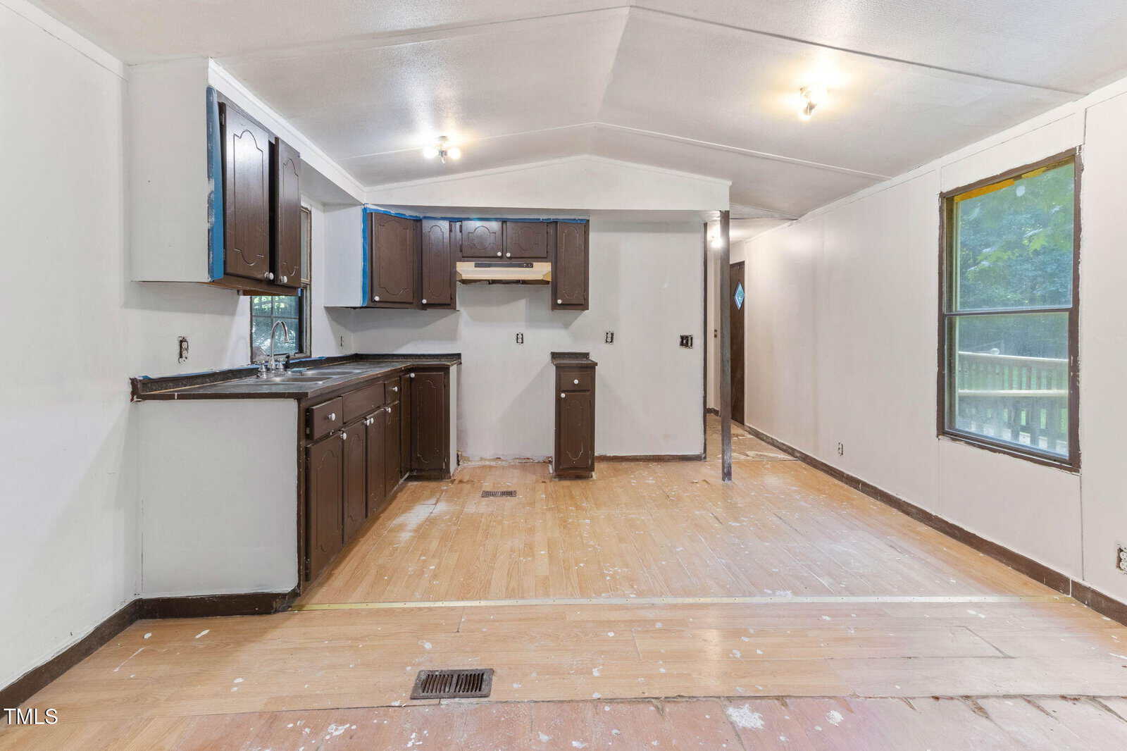 A partially renovated kitchen and living area with unfinished wood floors, brown cabinets, light gray walls, exposed outlets, and untrimmed windows. Some construction materials and paint marks are visible.