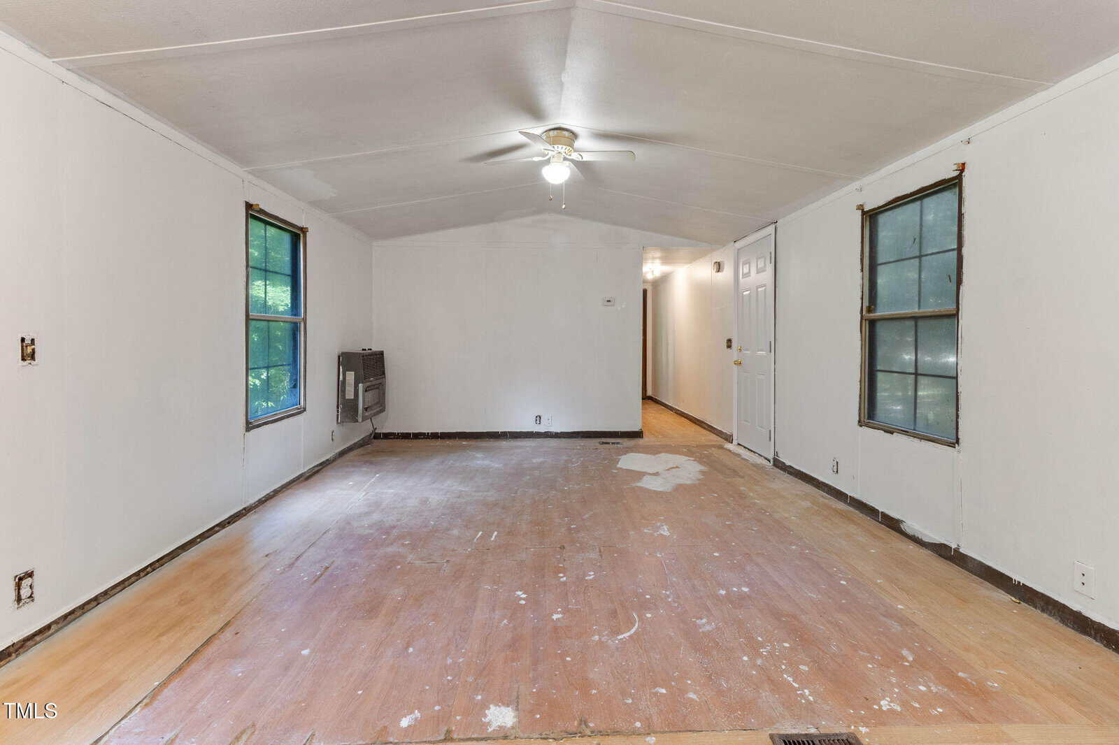 Empty room with unfinished floors, white walls, two windows on each side, ceiling fan with light, and a hallway leading to white doors in the background.