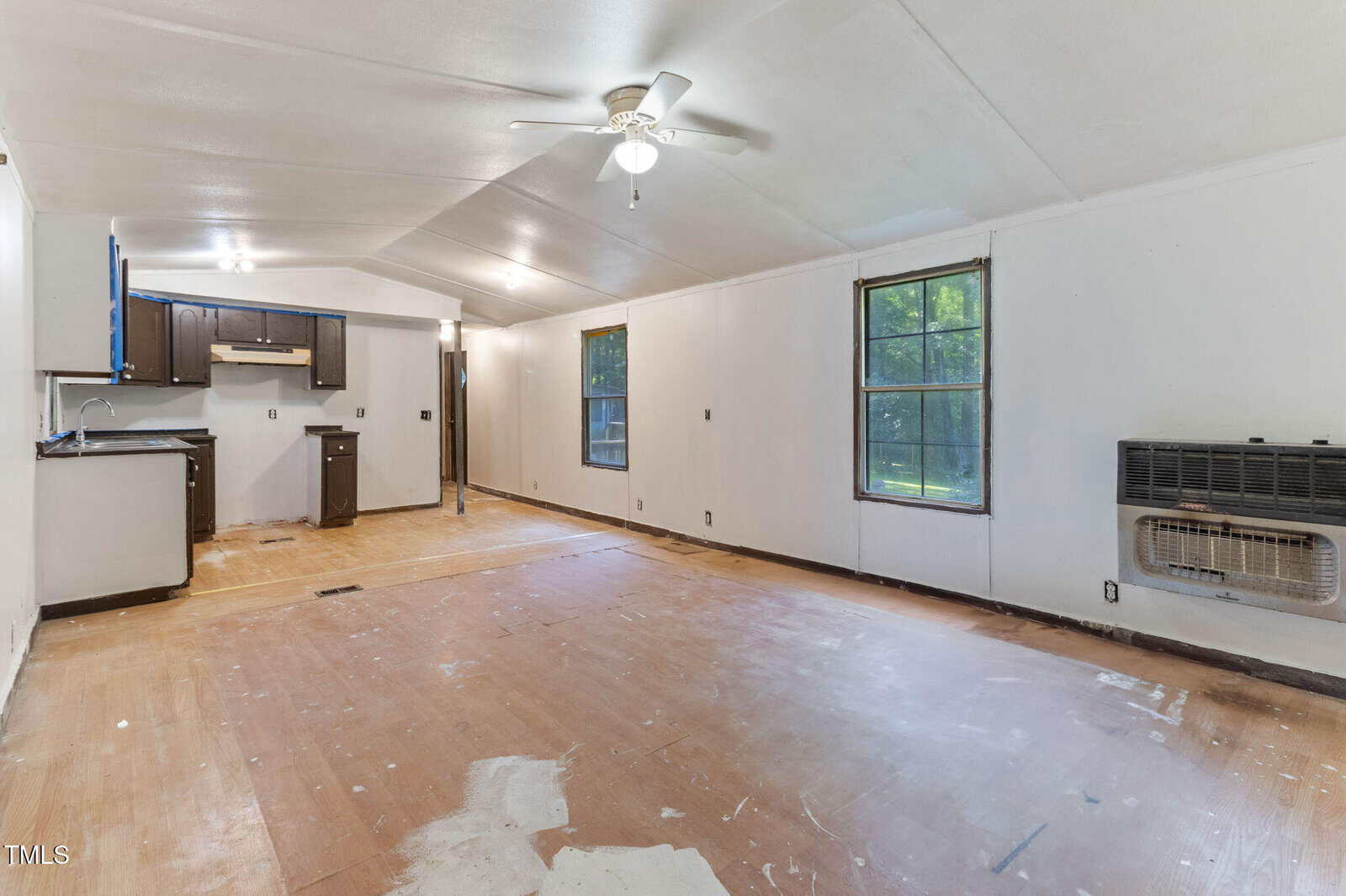 Unfurnished room with white walls, wood floors, two windows, a ceiling fan, wall heater, and a small open kitchen with dark cabinets in the back corner. Natural light fills the space.