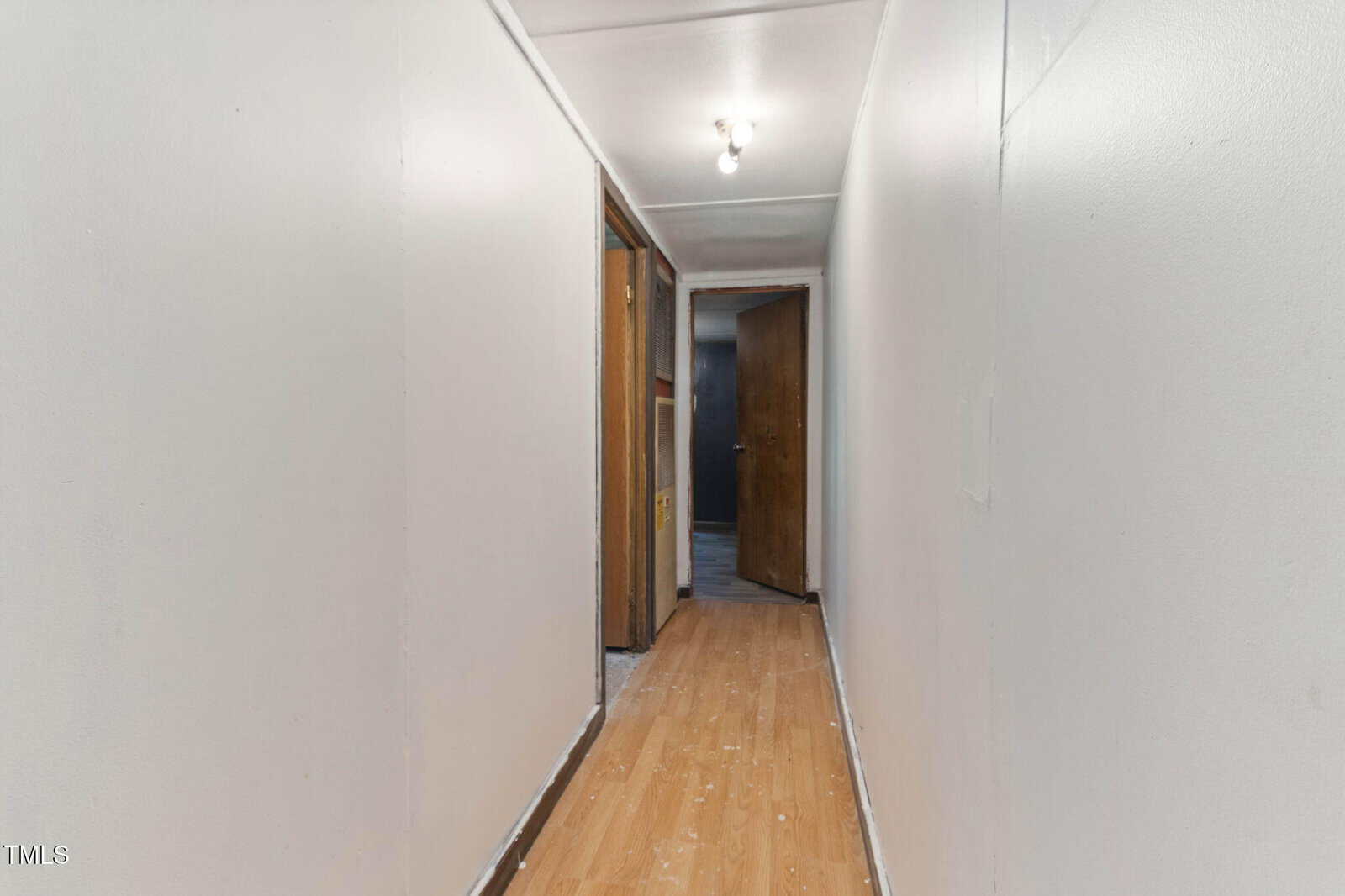A narrow hallway with white walls, a wooden floor, overhead lights, and an open brown door at the end leading to another room.