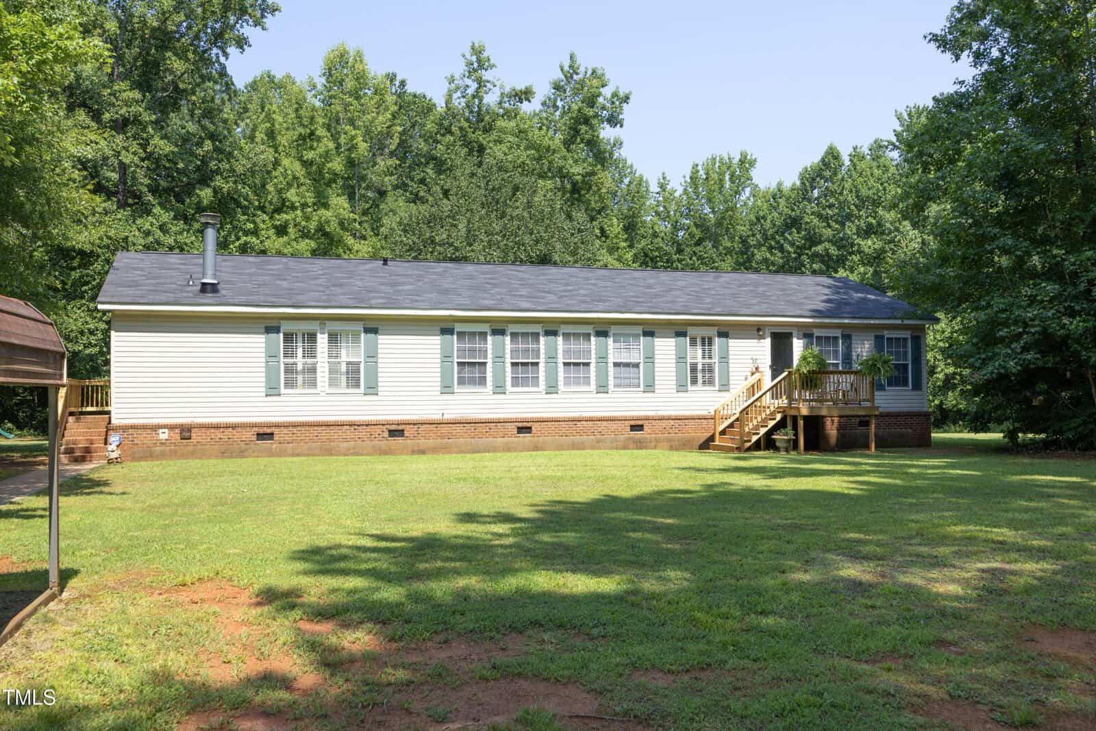 Single-story manufactured home with beige siding, numerous windows, a black shingle roof, and a small wooden porch. The house is surrounded by a grassy yard and backed by dense green trees under a clear sky.