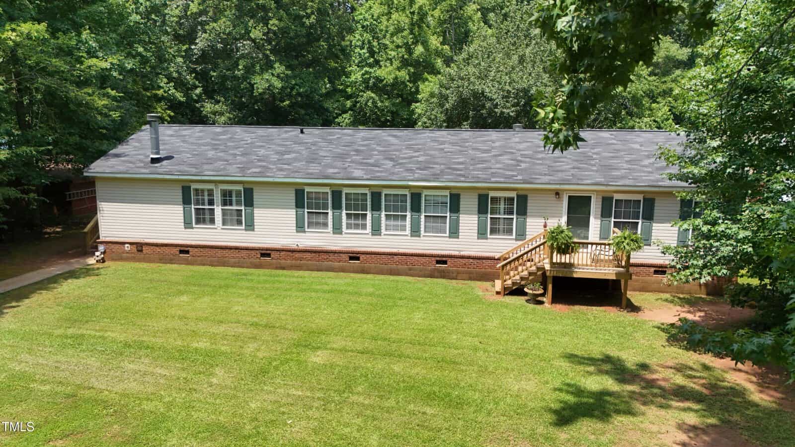 A single-story house with white siding, a brick foundation, multiple windows, and a small wooden front porch sits amid a spacious, neatly mowed lawn and surrounded by trees.