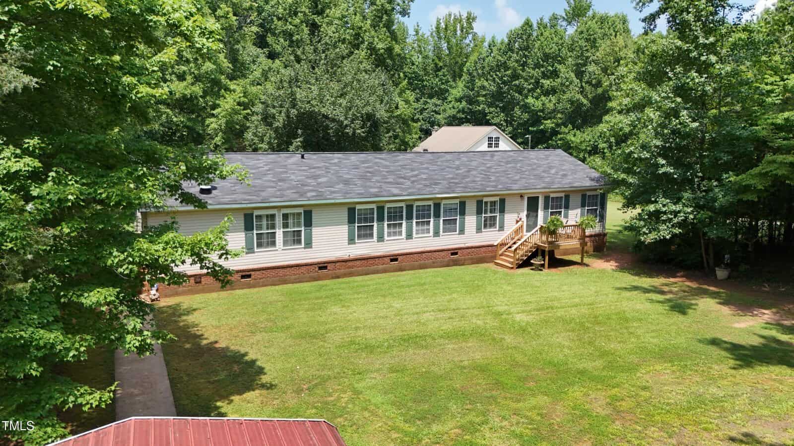 Single-story house with white siding and black roof, set on a grassy lawn. A wooden staircase leads to the front entrance. Surrounded by trees, with another building visible in the background.