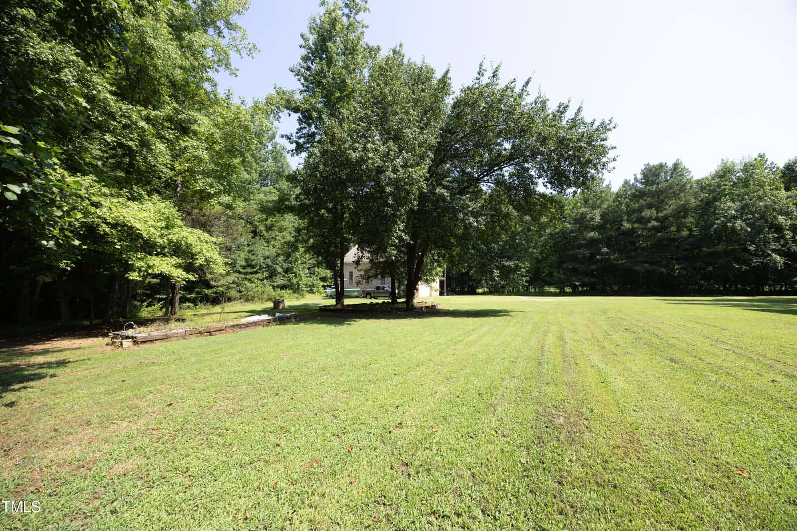 A large grassy yard bordered by trees, with a small white house partially visible in the center, shaded by a few large trees under a clear blue sky.