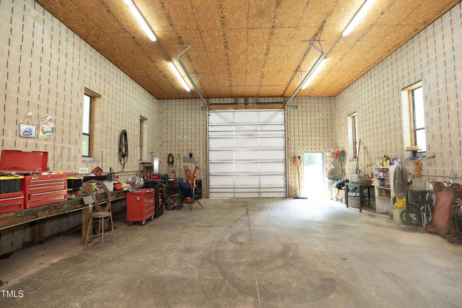 A spacious, well-lit garage with a high ceiling, concrete floor, and large overhead door. Workbenches, toolboxes, and various tools line the walls, creating a functional workshop space.