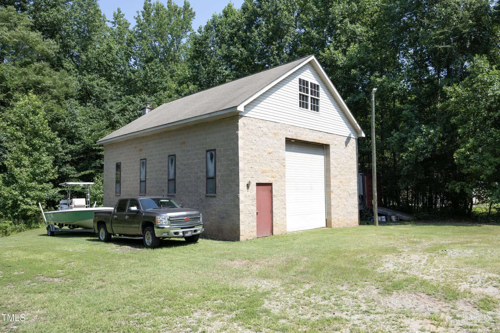 A tan brick garage with a white roll-up door and a red side door sits on a grassy yard. A silver pickup truck and a boat on a trailer are parked beside the building, with trees in the background.