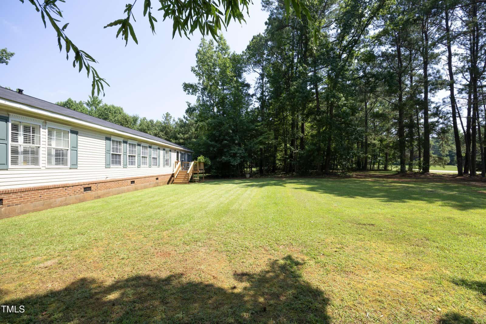 Single-story house with white siding and multiple windows on the left, bordered by a well-maintained grassy yard and surrounded by tall trees under a clear sky. Wooden steps lead to a door at the back of the house.