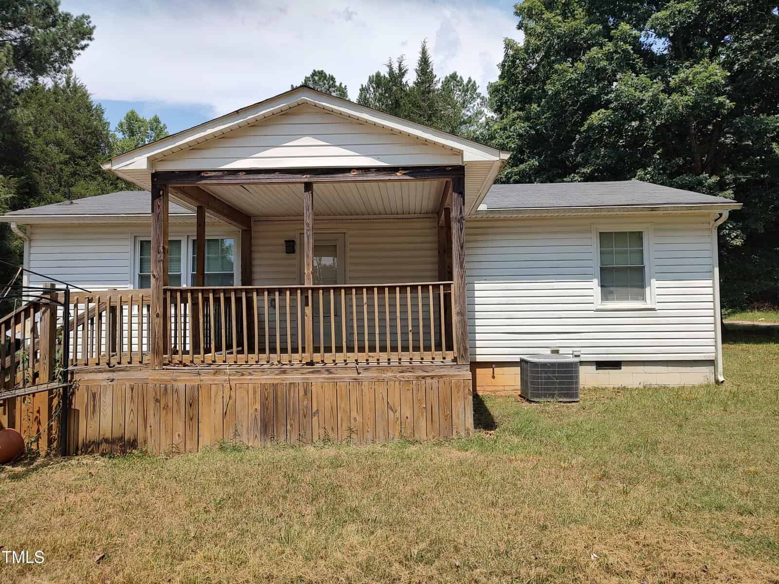 Single-story white house with a covered front porch featuring wooden railings and steps. There is an air conditioning unit on the right side and a grassy yard in the foreground with trees in the background.