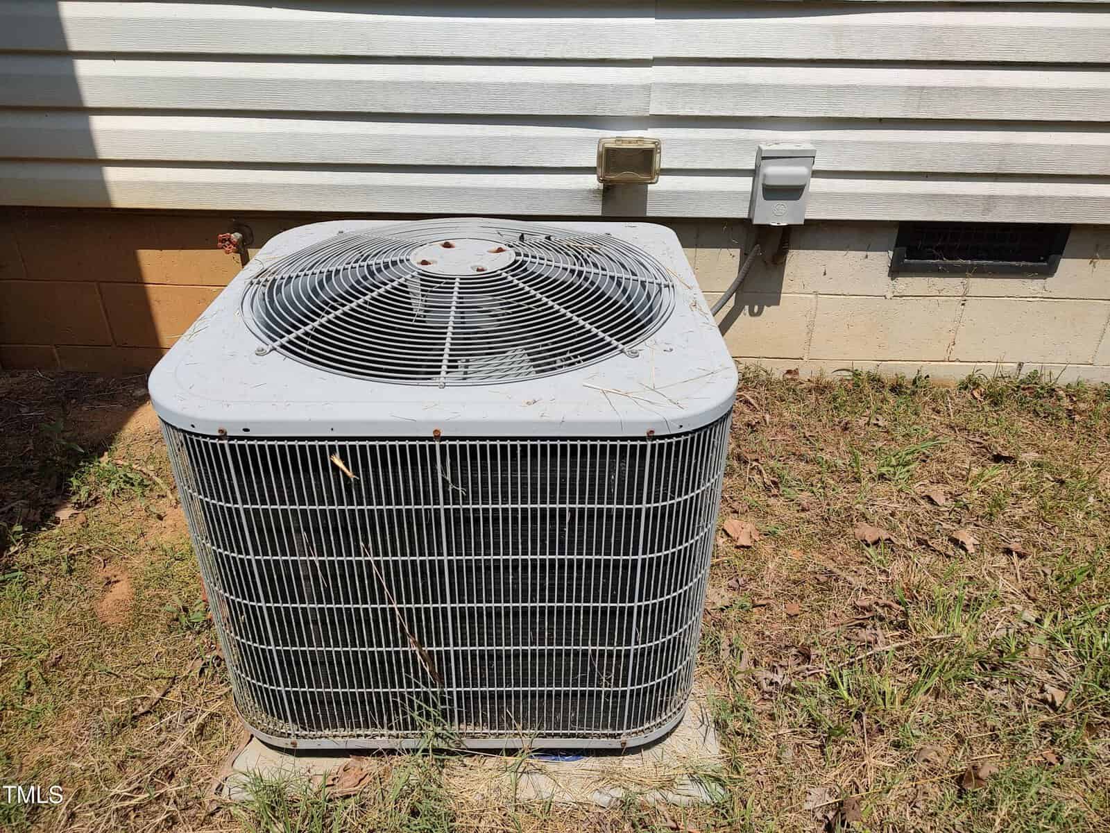 A large outdoor air conditioning unit sits on a concrete pad next to the exterior wall of a house, with electrical outlets and a vent visible on the wall behind it. Grass and dirt cover the ground around the unit.