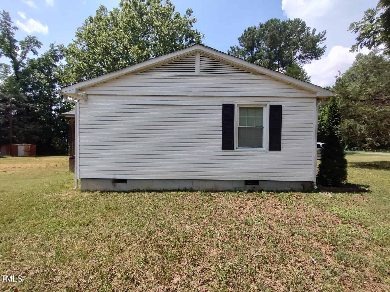 A single-story house with white vinyl siding, a single window with black shutters, and a grassy yard. Large trees are visible in the background under a partly cloudy sky.