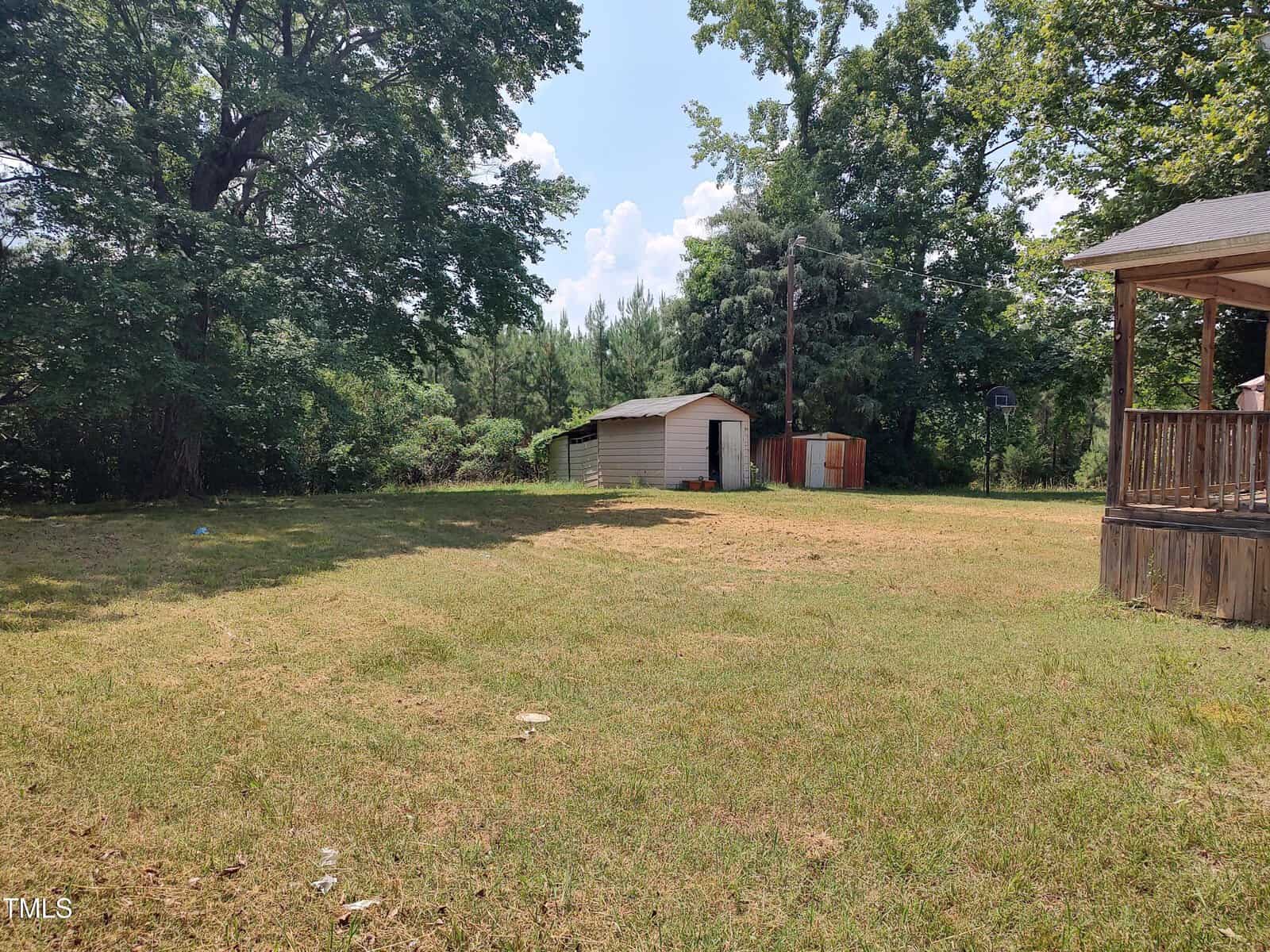 A grassy backyard with a few large trees, a small white shed, a red storage structure, and a wooden deck attached to a house on the right side. The sky is mostly clear with some clouds.