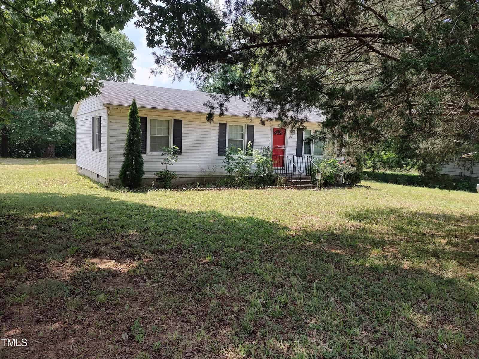 A single-story white house with a red front door and small porch sits in a grassy yard at 1161 Morgan Road, Henderson, partially shaded by large trees. Shrubs and plants border the front of the house.