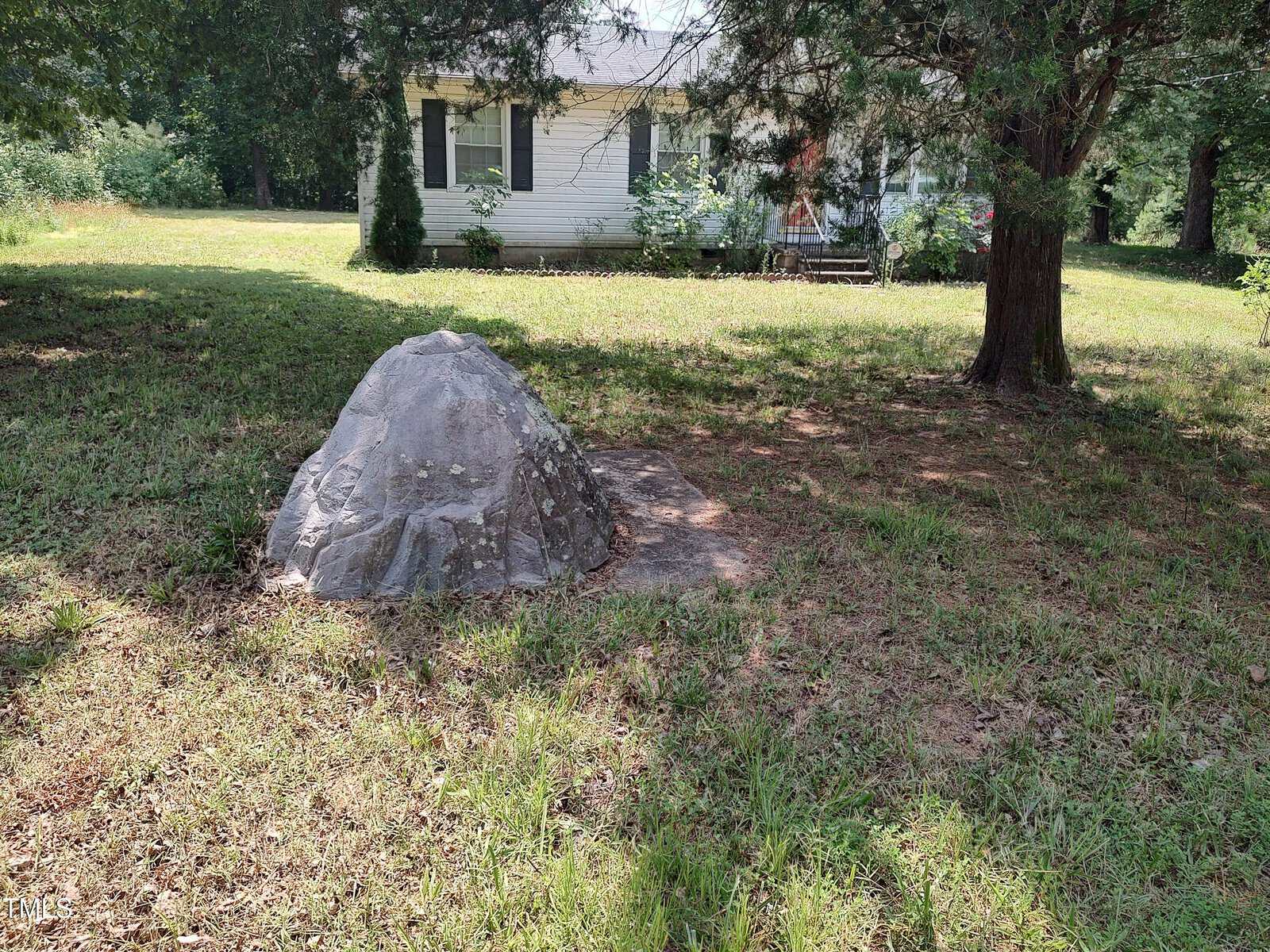 A large artificial rock covers something in a grassy yard with patches of sunlight, with a white house, bushes, and trees in the background.