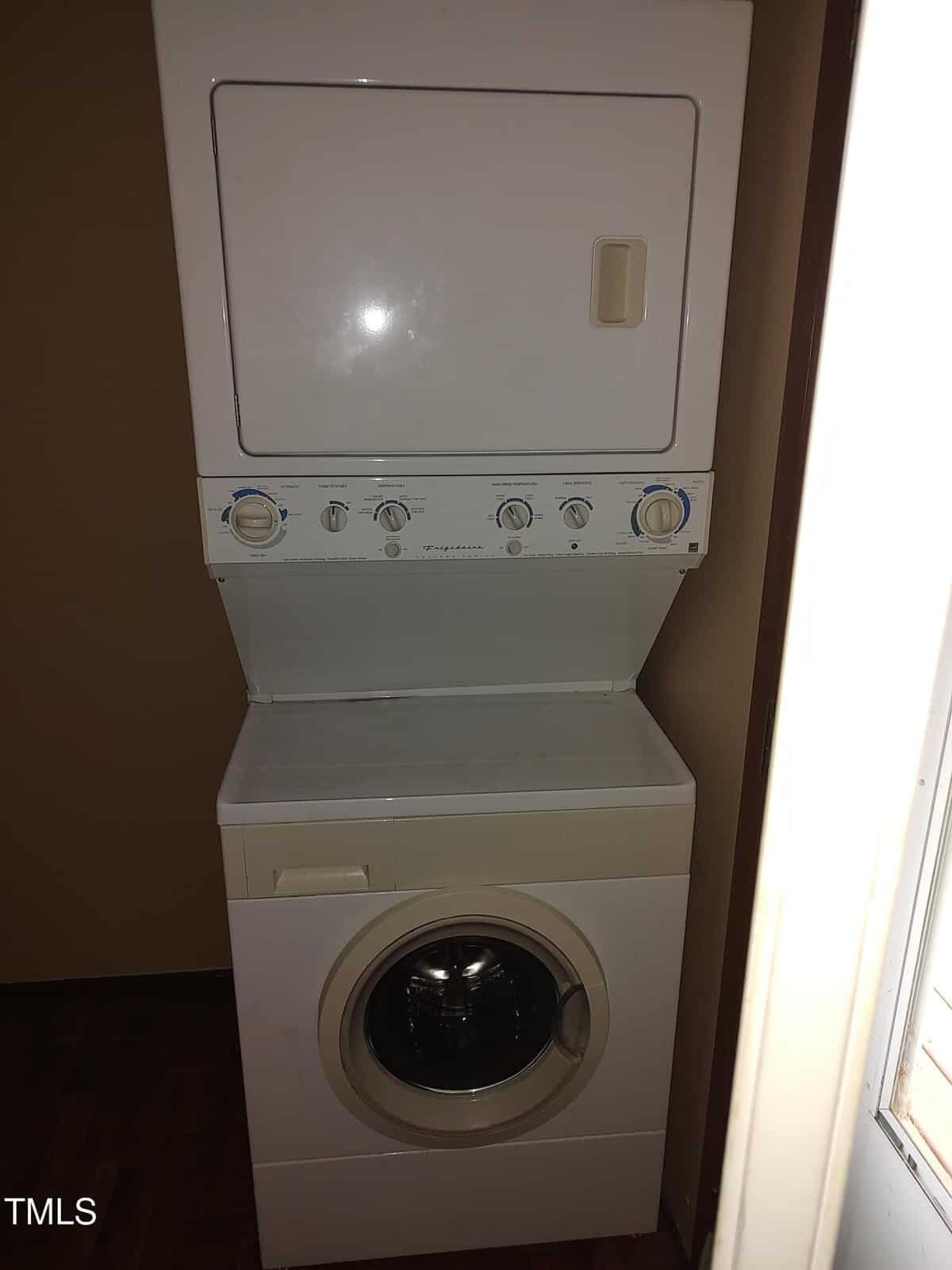 A stacked washer and dryer unit is placed in a small, dimly lit laundry area with brown walls and a window on the right.