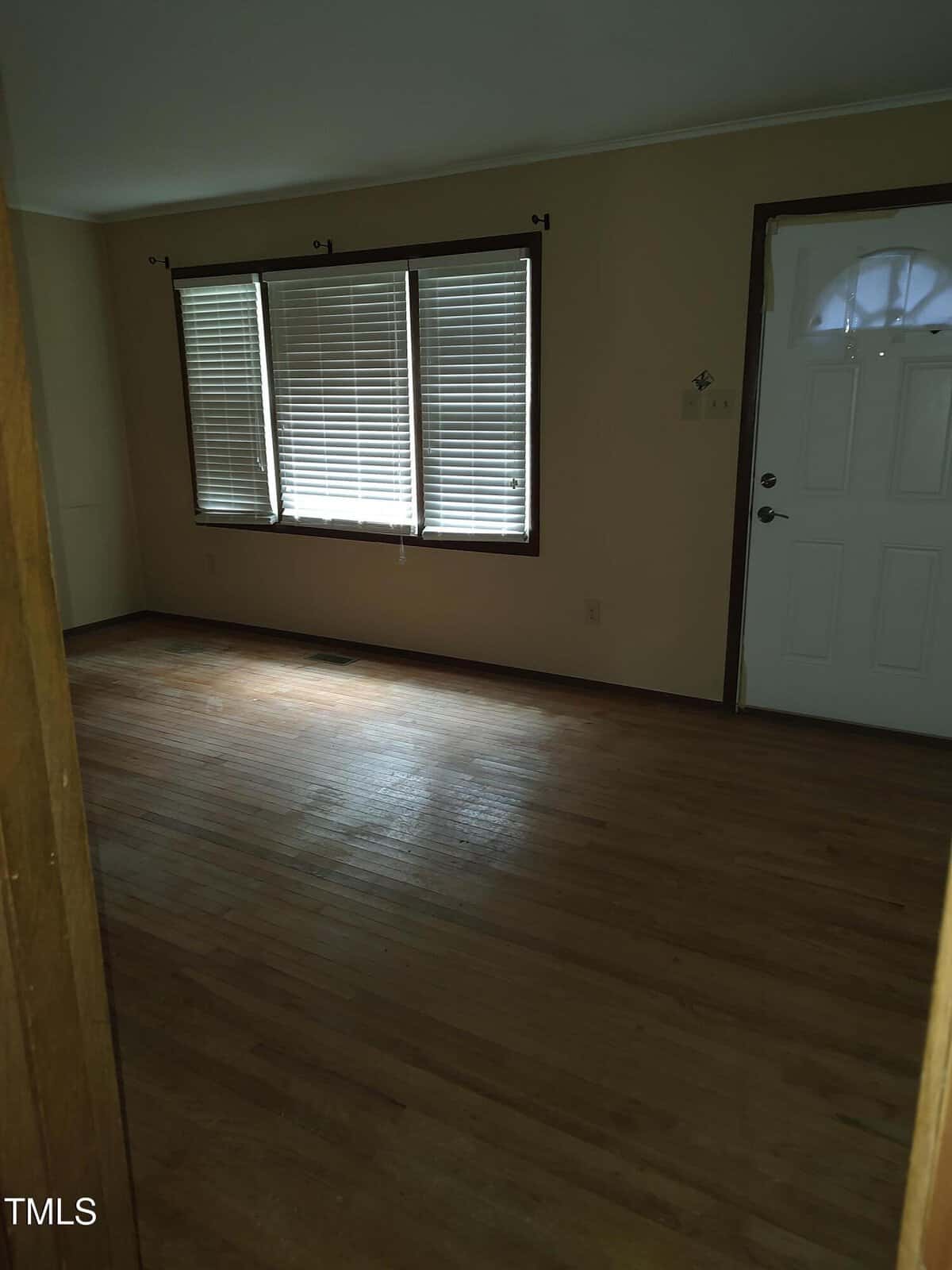 An empty room with hardwood floors, beige walls, three windows with blinds, and a white front door. Natural light enters through the windows, creating shadows on the floor.