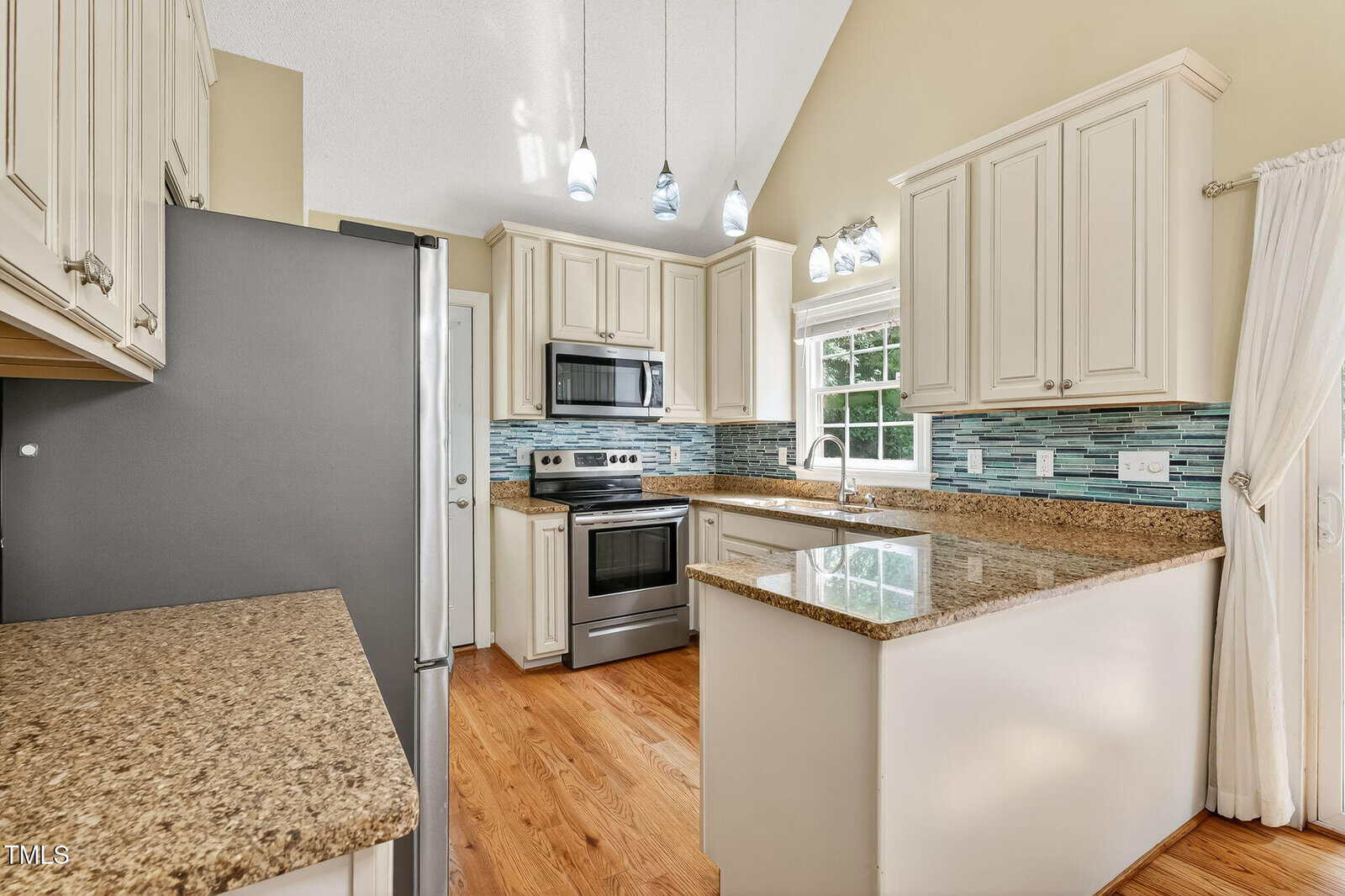 Modern kitchen with white cabinets, granite countertops, stainless steel appliances, blue mosaic tile backsplash, and hardwood floors. Pendant lights hang from a high ceiling above the peninsula countertop. Sunlight enters through a window.
