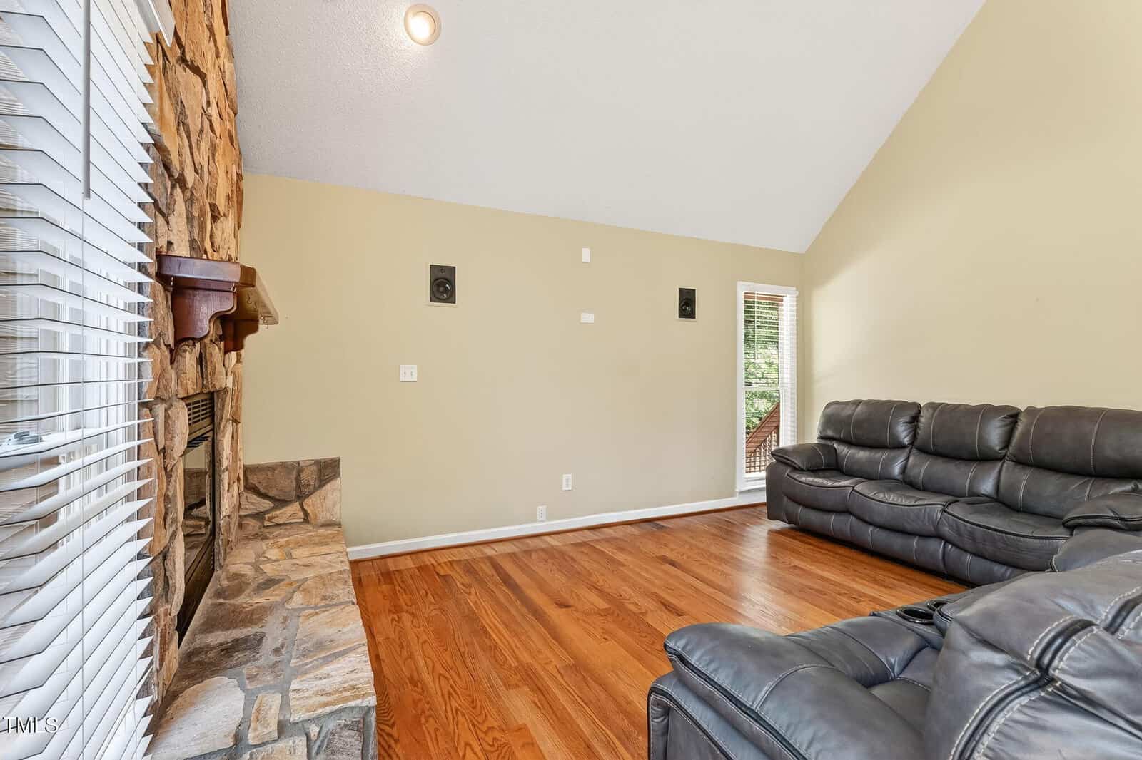 Living room with hardwood floors, stone fireplace on the left, beige walls, two black leather sofas, wall speakers, and a glass door leading outside. Natural light comes in through large windows on the left.