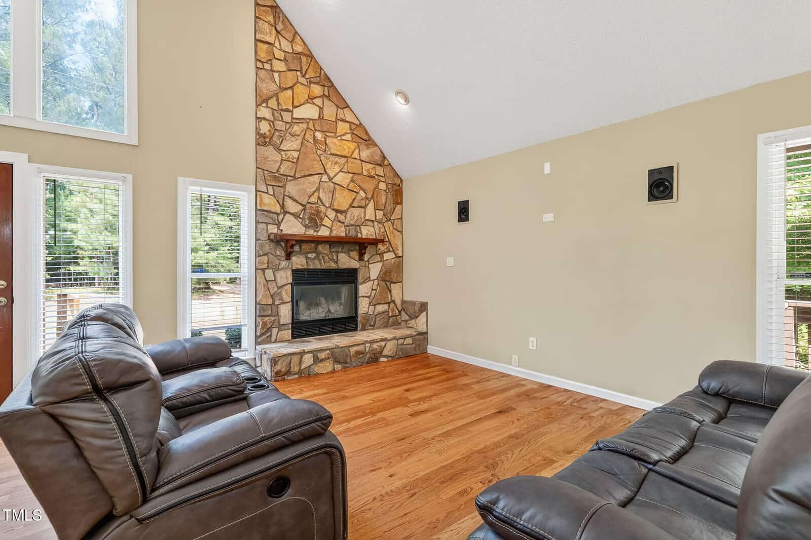 Living room with tan walls, hardwood floors, two brown leather sofas, a stone fireplace with a mantel, high ceiling, large windows, and wall-mounted speakers. Natural light fills the space through the windows.