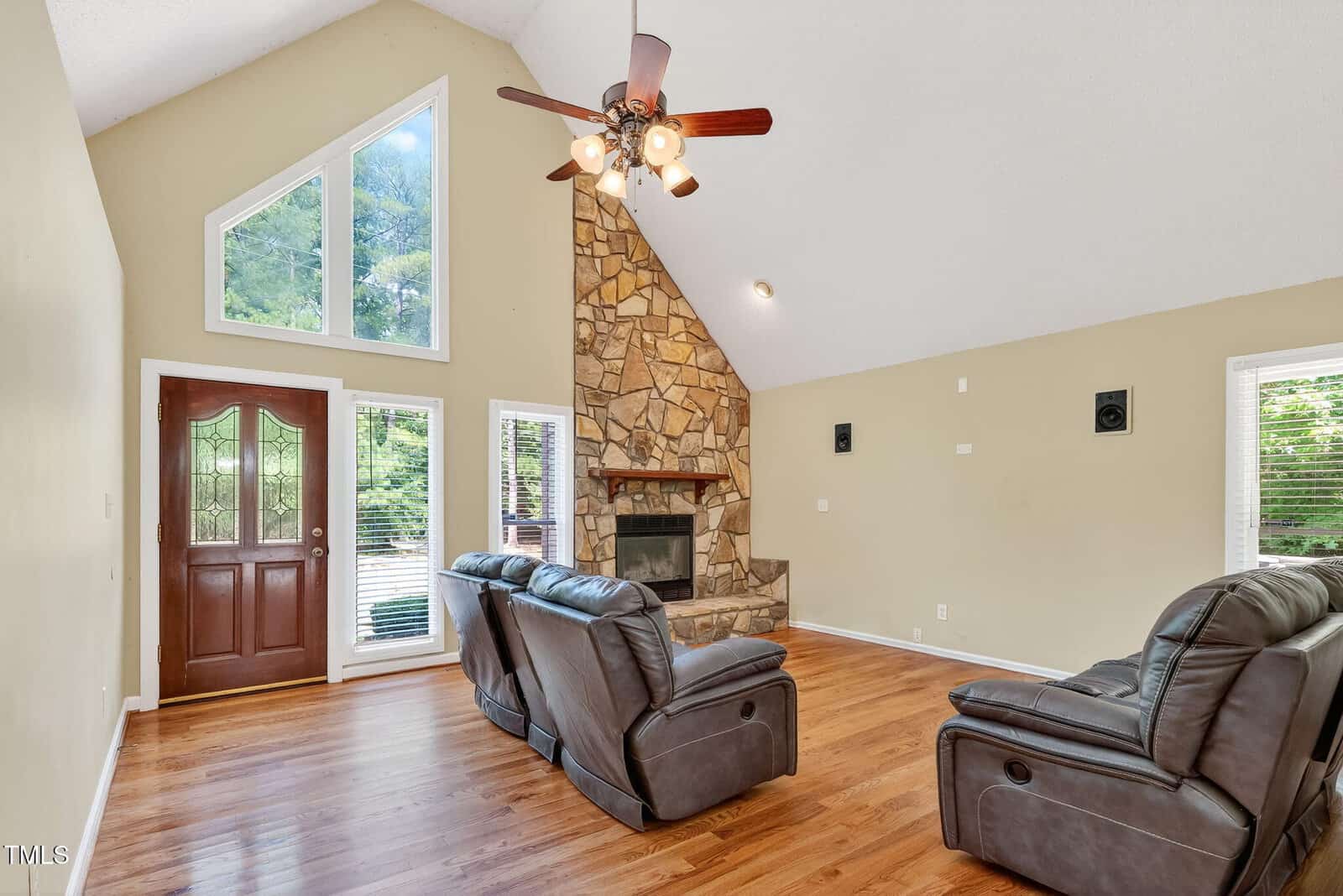 A spacious living room with vaulted ceiling, hardwood floors, two leather recliners, a stone fireplace, large triangular windows, and a wooden front door with glass panels.