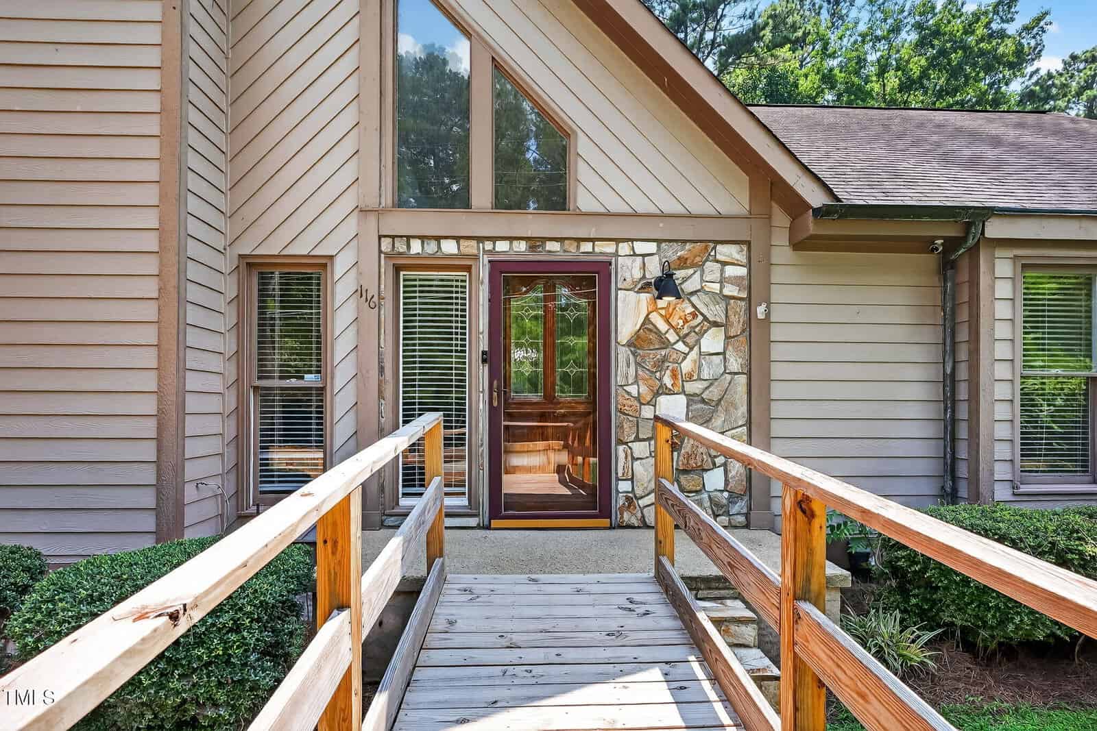 A wooden ramp with railings leads to the glass front door of a modern house with beige siding, stone accents, large windows, and neatly trimmed shrubs.