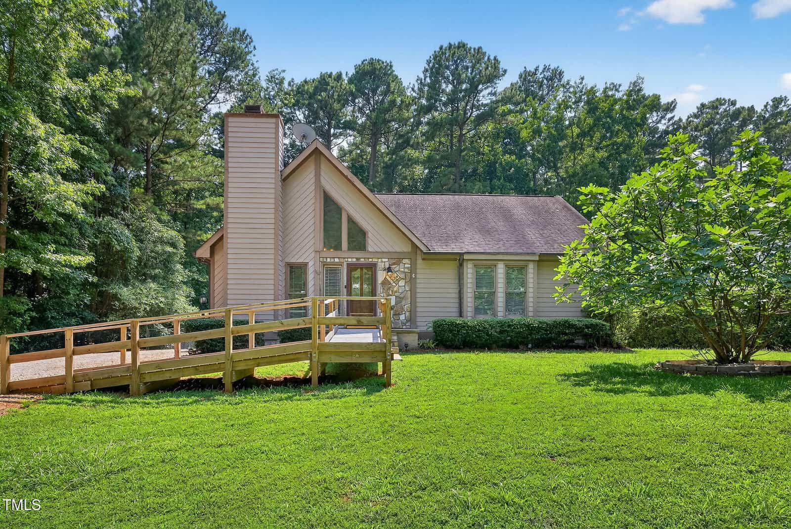 A single-story house with a steep roof and large front windows, surrounded by tall trees. A wooden wheelchair ramp leads to the front door across a well-maintained green lawn under a blue sky.