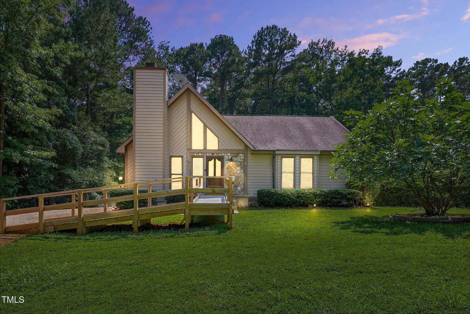 A single-story house with a peaked roof and large windows glows warmly at dusk. Surrounded by lush green lawn and trees, the house features a wooden ramp leading to the front entrance.