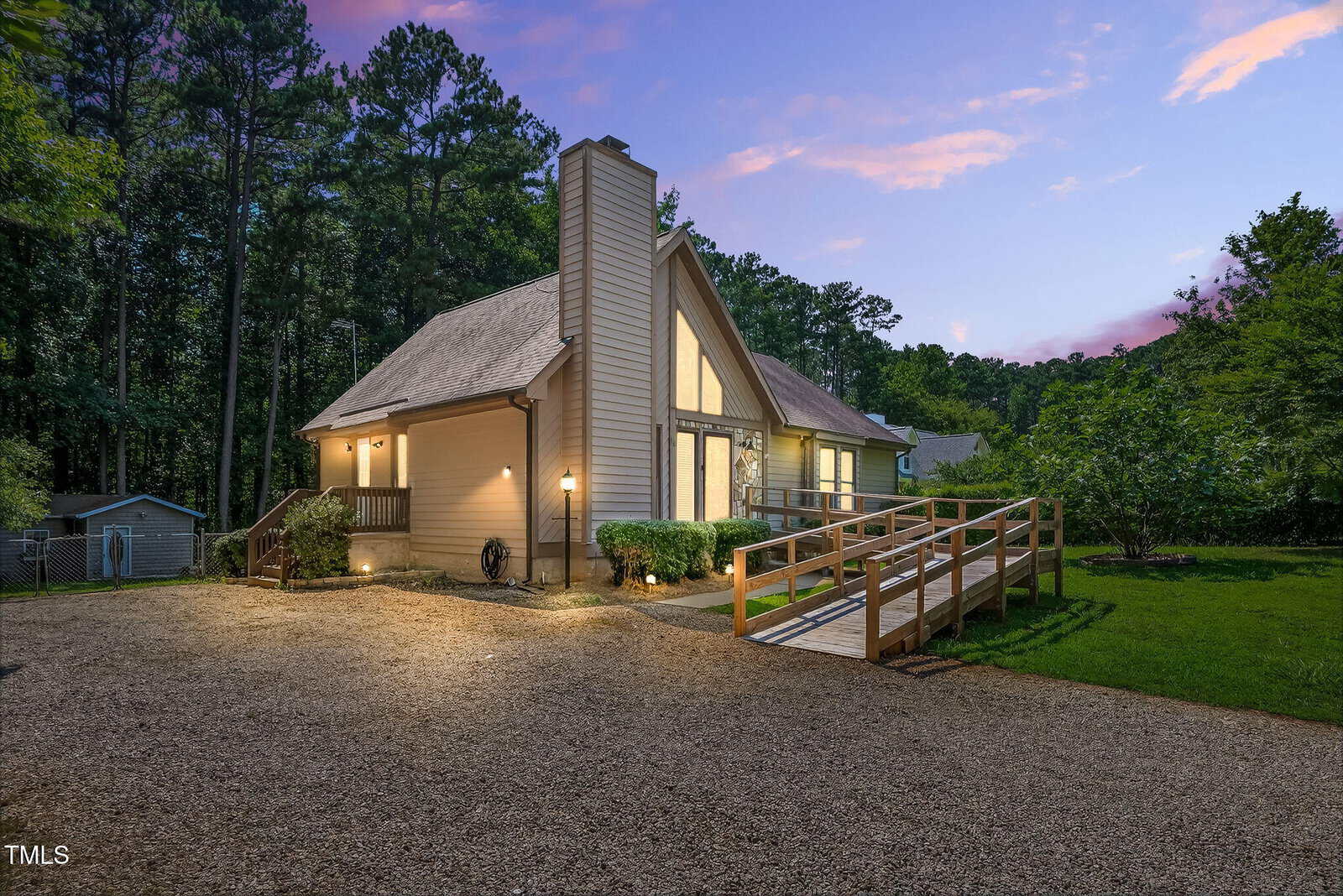 A modern, light-colored house with large windows and a steep roof stands at dusk. The home has a wooden ramp leading to the front door, exterior lights, and is surrounded by trees and a gravel driveway.