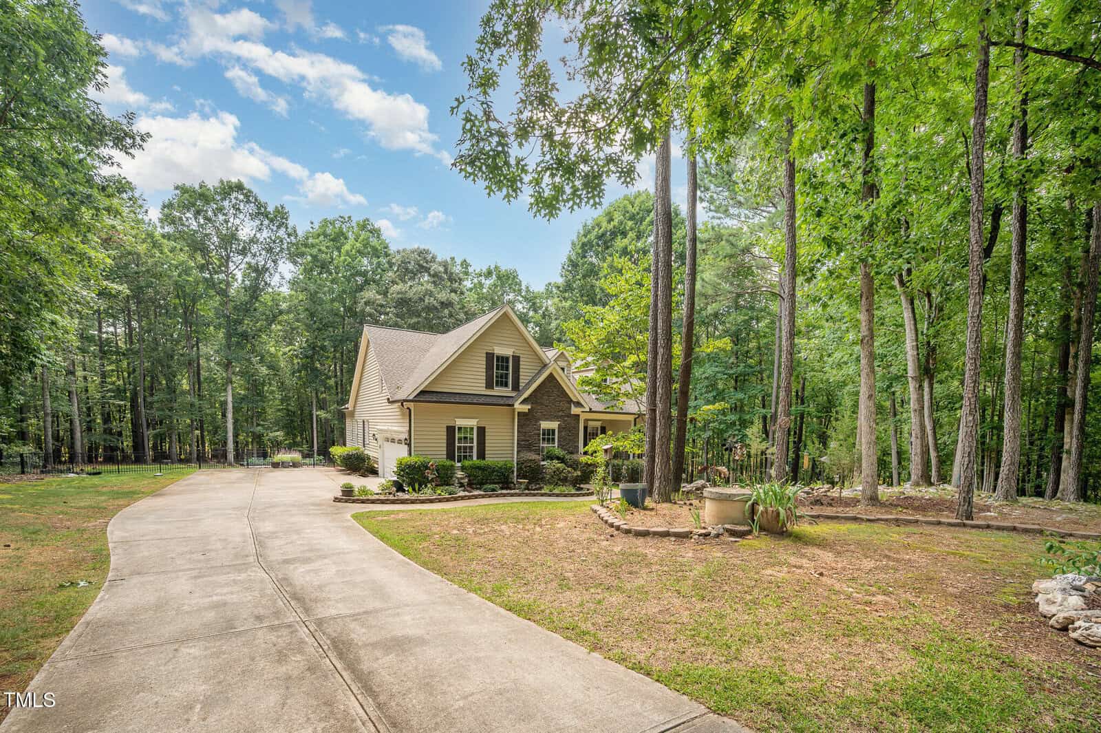 A beige, single-story house with a gabled roof sits among tall trees at 1513 Anterra Drive in Wake Forest. A wide concrete driveway curves to the entrance, with landscaping, potted plants near the front, and a partly cloudy sky above.