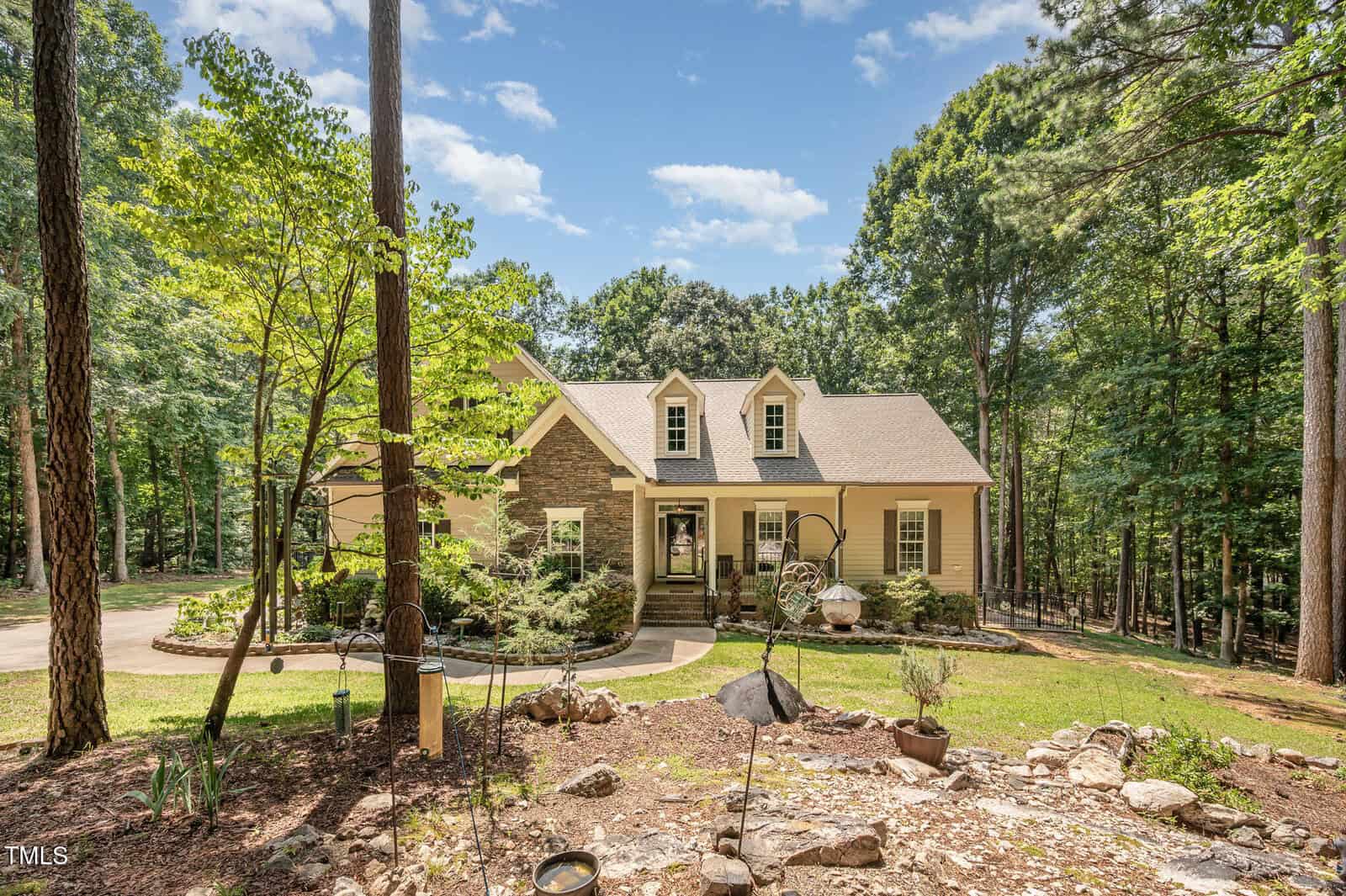 A two-story house with a stone and beige exterior sits among tall trees. The house has three dormer windows, a covered porch, and landscaped gardens in front. Sunlight filters through the surrounding forest.