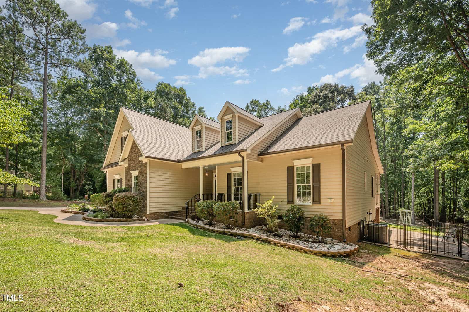 A beige two-story house with stone accents, gabled roof, covered front porch, and white railings, surrounded by trees and landscaped with bushes and small rocks under a blue sky with scattered clouds.